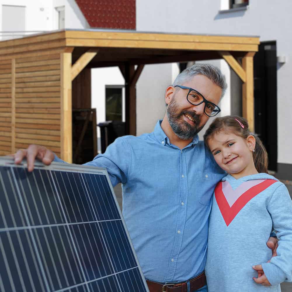 Vater mit Kind, beide lächeln, hält ein Solarmodul. Im Hintergrund ein Holzcarport.