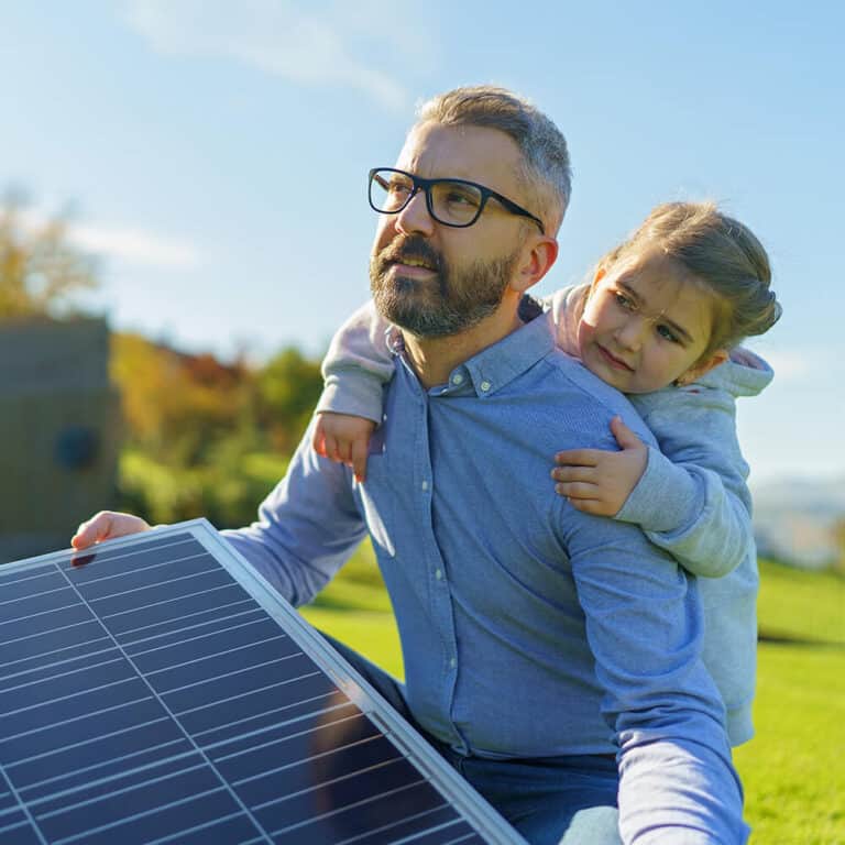 Mann mit Kind blickt in die Ferne und hält ein Solarmodul. Im Hintergrund der Garten seines Hauses.